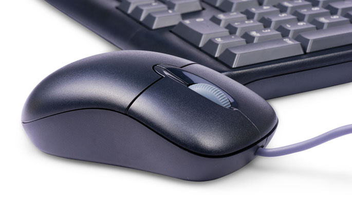 A dark grey computer mouse, with a keyboard in the background. Both are on a white surface.