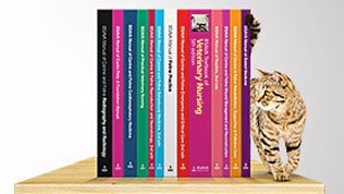 A cat next to a collection of BSAVA manuals sitting upright on a table, book spines showing