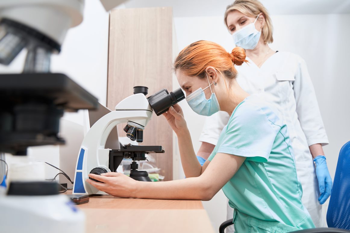 A student is observed and guided by her teacher, as she looks at a sample through a microscope in a laboratory